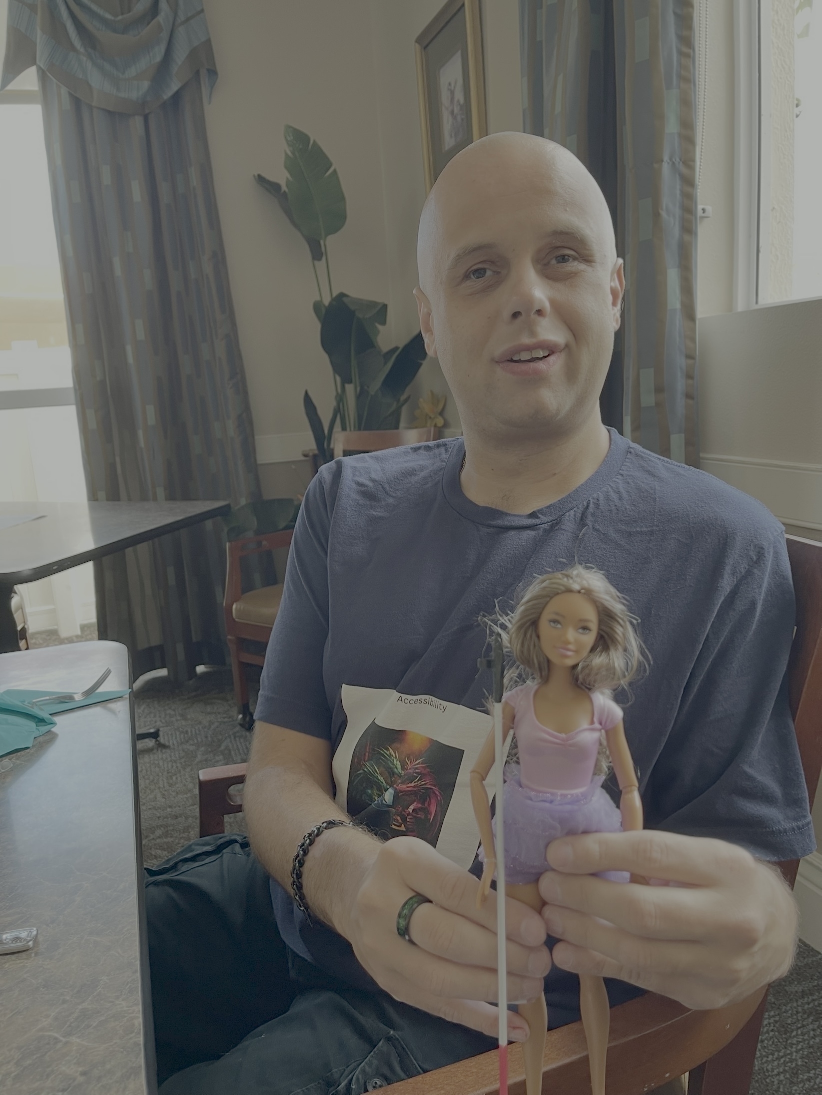 The image shows a person sitting indoors, holding a Blind Barbie® doll. The person is bald and wearing a dark blue T-shirt with a graphic design on it. They are smiling slightly and looking towards the camera. The Blind Barbie® doll they are holding has long brown hair and is dressed in a pink top and a purple skirt. The doll is also holding a small white cane, similar to those used by visually impaired individuals. In the background, there is a table with some items on it, a large plant, and a window with curtains. The setting appears to be a casual indoor environment, possibly a dining or living area.