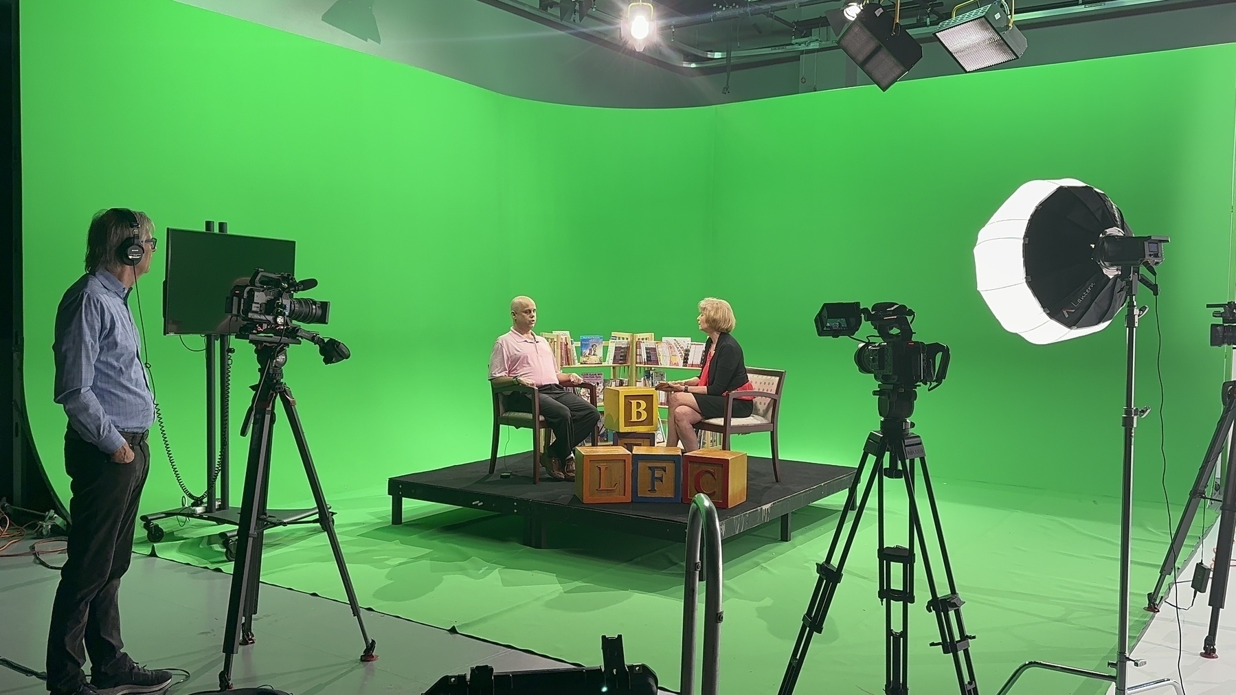 Auto-generated description: A green screen studio setup captures an interview between two people seated on a platform, surrounded by cameras and lighting equipment.