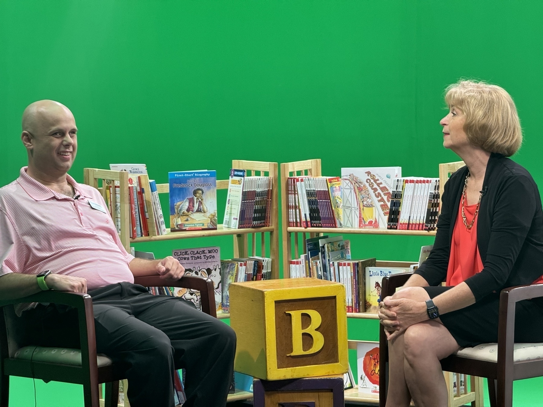 Auto-generated description: Two people are sitting and conversing in front of a green screen with several bookshelves filled with books between them.