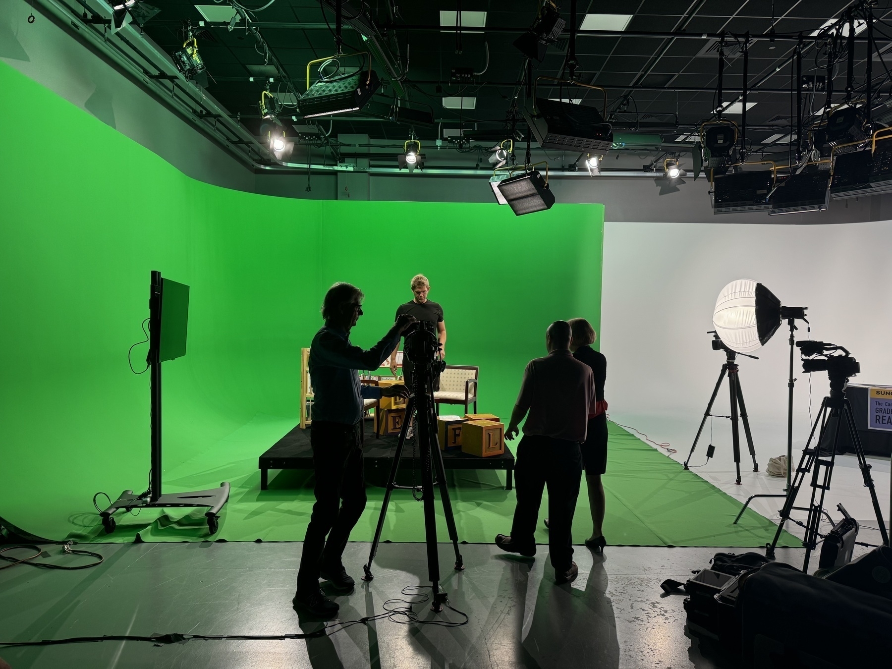 Auto-generated description: A group of people is working with cameras and equipment in a studio with a green screen and stage setup.