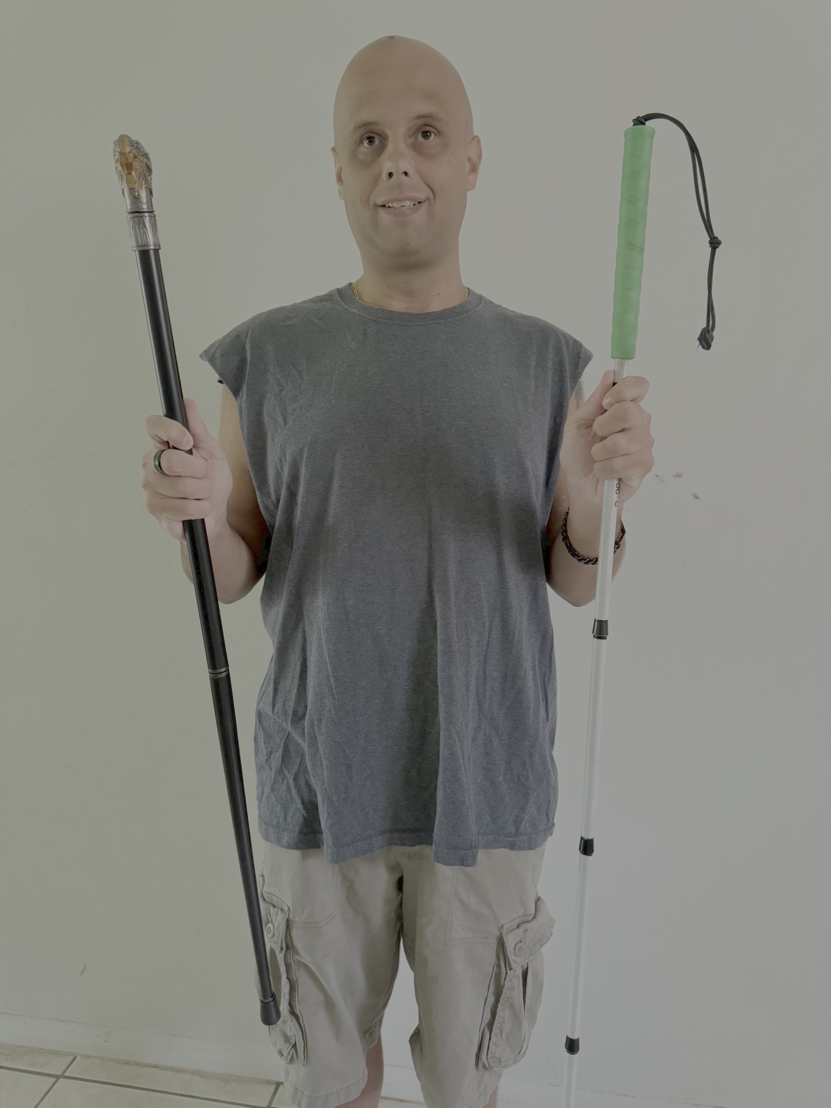 The image shows a person standing indoors against a plain light-colored wall. They are bald and smiling. They are wearing a sleeveless gray t-shirt and beige cargo shorts. In their right hand, they are holding a black cane with a handle shaped like a dragon's head. In their left hand, they are holding a white cane with a green handle and a black wrist strap. They are also wearing a medical alert bracelet on their left wrist.