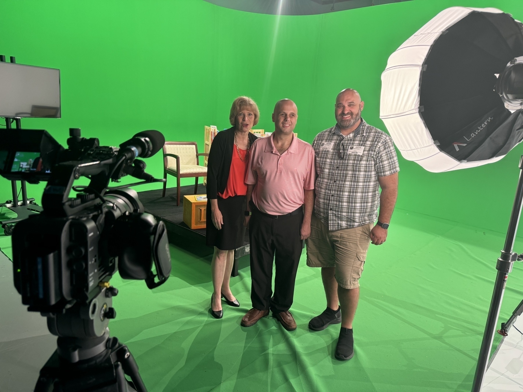 Auto-generated description: Three people are standing and smiling in a green screen studio, surrounded by filming equipment and lights.