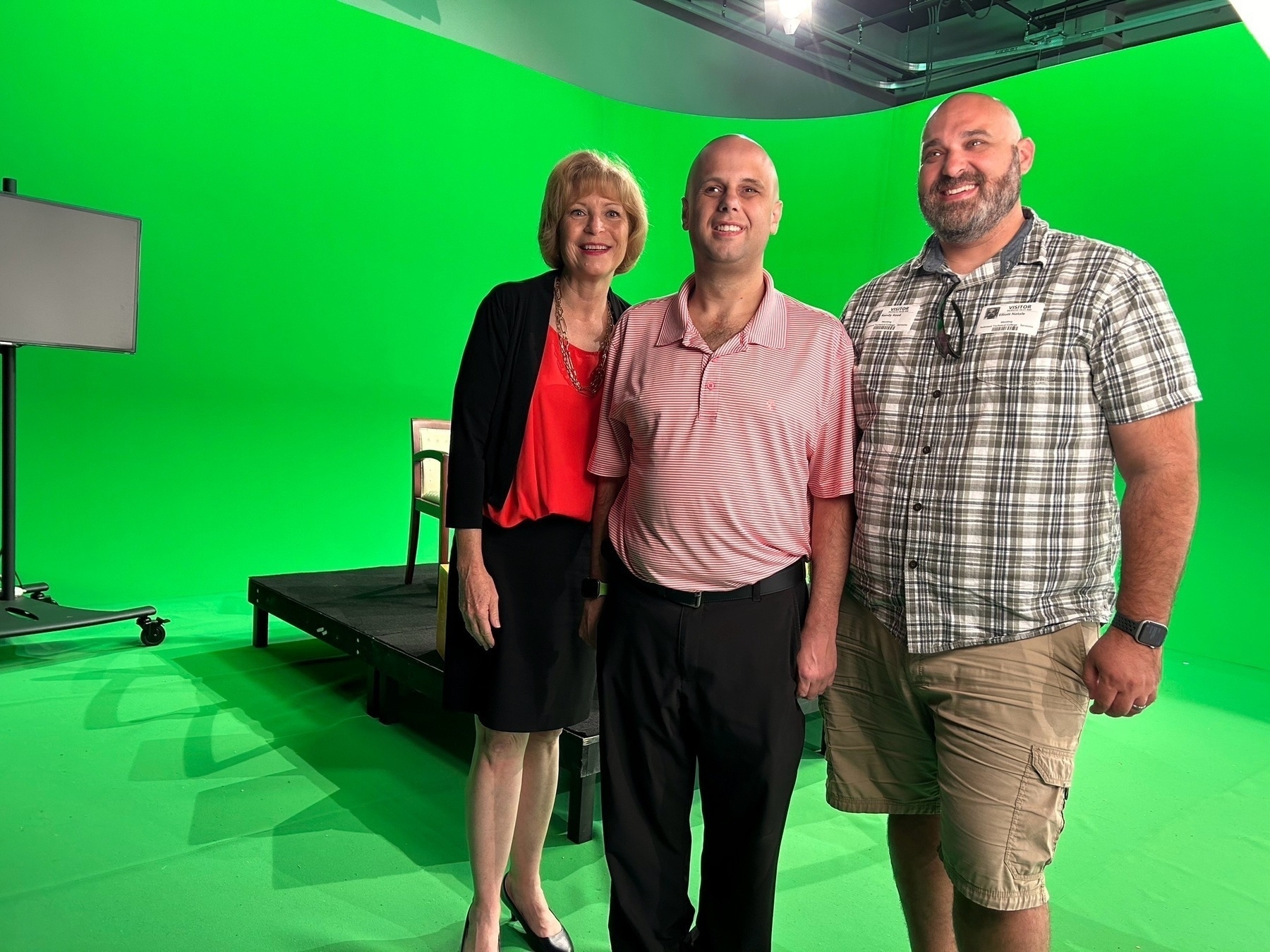 Auto-generated description: Three people are standing in front of a green screen on a set with stage equipment visible.